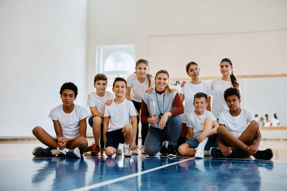 Portrait Of Happy Sports Teacher With Group Of Kids At School Gym Looking At Camera.